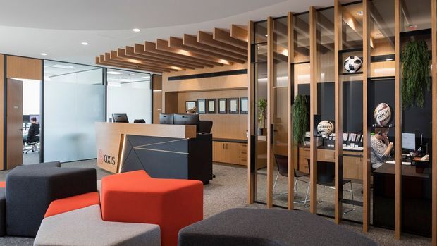 Contemporary commercial office reception and lounge area with a branded reception desk, modular upholstered seating, glass-partitioned workspaces, timber cabinetry, and a dramatic open timber-slat ceiling feature. A tall timber-framed open shelving divider displays sports memorabilia and integrated greenery, creating separation between the entry lounge and office area.