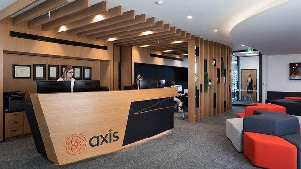 A contemporary commercial reception area with a large angled timber reception desk bearing the Axis logo, black front detailing, and integrated workstations. The space features extensive warm timber wall panelling, vertical timber slats forming a screened corridor, and a dramatic open timber batten ceiling feature. Framed certificates line the wall behind reception, while colourful modular lounge seating occupies the foreground. Glass doors and office areas are visible beyond the reception zone.