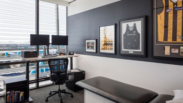 Compact, contemporary home office overlooking an outdoor sports venue. The room features a built-in desk with dual monitors, a black ergonomic office chair, dark carpet, a white storage unit, and a long black upholstered bench or daybed. A charcoal feature wall displays framed sports memorabilia, while large windows with horizontal blinds provide abundant daylight and an urban stadium view.
