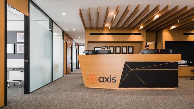 A contemporary commercial office reception area with a large angular timber and black reception desk bearing the Axis logo. The space features warm timber wall cladding, a dramatic exposed timber-slat ceiling, charcoal carpet, glass-partitioned offices, framed wall displays, recessed lighting and integrated workstations behind the desk.