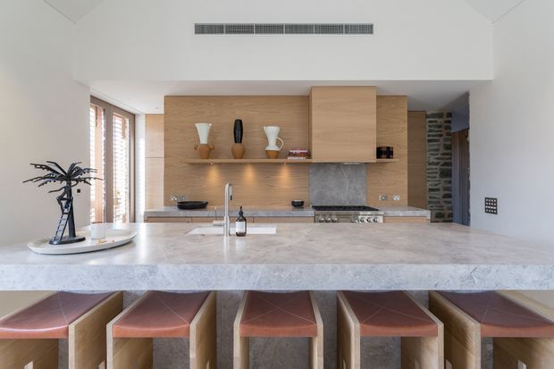 Bright contemporary kitchen viewed from the dining or living side, featuring a large pale stone island with integrated sink and seating for several people, light timber cabinetry and open shelving, a stone splashback, stainless steel cooking appliances, and warm wood-and-leather bar stools. A high angled ceiling with linear air vent and tall windows with timber shutters add architectural definition.