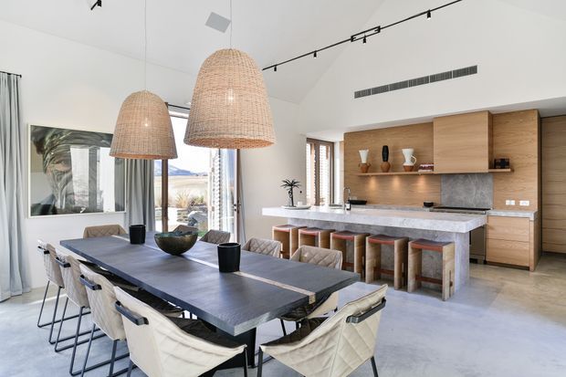 Bright open-plan dining and kitchen interior with a long dark dining table, upholstered dining chairs, woven pendant lights, and a large stone kitchen island. The kitchen features warm timber cabinetry, open shelving, a grey stone splashback, and integrated appliances. Floor-to-ceiling glazing and a high sloped ceiling create an airy, contemporary space.
