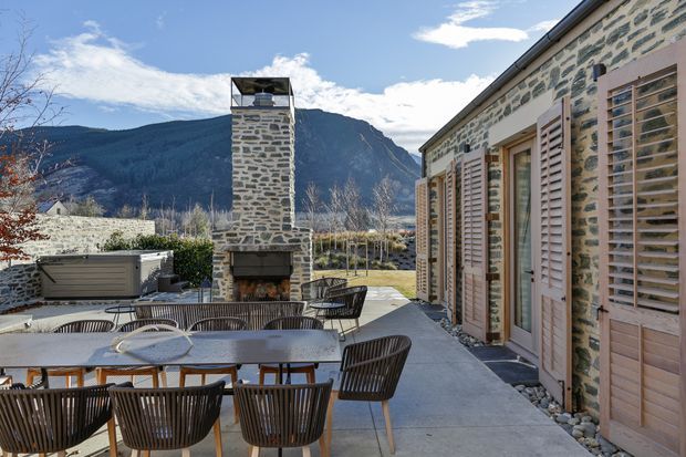 A sunlit outdoor dining terrace beside a stone-clad building, furnished with a long dining table and dark woven chairs. A substantial open fireplace with a tall stone chimney forms a focal point, while timber shutters and glazed doors line the building facade. Mountainous terrain is visible beyond the property.