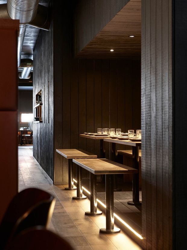A narrow, moody restaurant dining area with dark vertically panelled walls, timber ceiling slats, exposed industrial ductwork, and built-in bar-height communal tables. Warm linear lighting runs along the base of the tables, highlighting the dark metal pedestal supports and wood tabletops. A corridor leads toward a brighter dining area in the background.