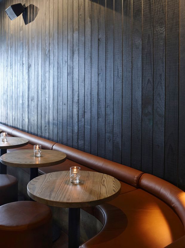 A dimly lit hospitality seating area with round timber tables, integrated brown leather banquette seating, matching leather stools, and small candle lights. The backdrop is a full-height wall clad in vertically arranged dark timber boards, with a black wall-mounted light fixture adding a subtle accent.
