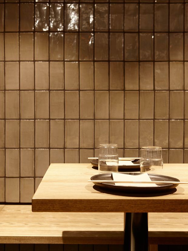 Close-up of a warm, intimate dining setting with a light timber table, dark pedestal base, ceramic plates, drinking glasses, and folded napkins. The backdrop is a full-height grid of glossy, irregular brown ceramic tiles, with matching timber flooring and bench-like seating visible below.