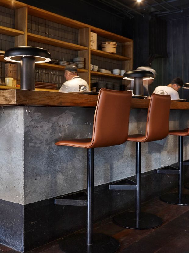A moody commercial bar or restaurant interior featuring a long counter with a textured dark grey concrete or plaster front, a warm timber countertop, and tall brown leather bar stools on black metal pedestals. Open timber shelving behind the counter displays stacked dishes and pottery, with dark tiled walls, black ceilings, and distinctive pendant lamps creating an industrial, refined atmosphere.