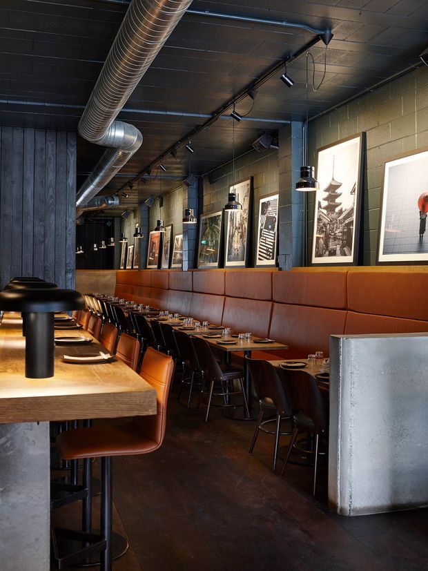 A narrow industrial-style restaurant or bar interior with exposed ductwork, dark painted ceiling, track lighting, concrete block walls, a long tan leather banquette, timber dining tables, leather dining chairs and stools, and framed artwork along the wall.