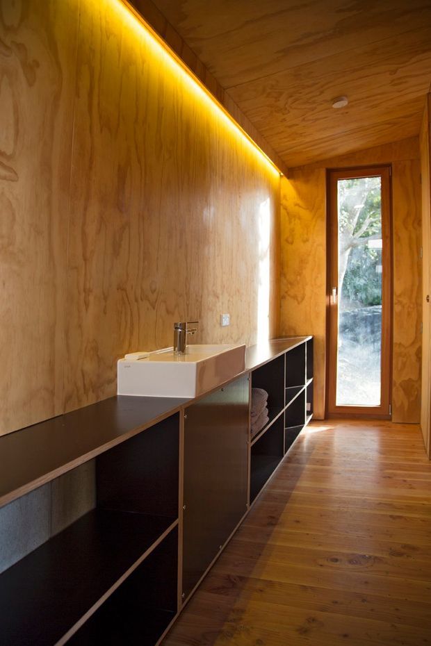 A narrow, warm-toned bathroom or washroom lined throughout with natural plywood, including the walls and ceiling. A long dark built-in vanity with open shelving runs along one side and supports a white rectangular vessel basin with a chrome tap. Integrated LED strip lighting highlights the upper wall, while a tall timber-framed glazed door brings in daylight and views of greenery. The timber floor and dark joinery create a compact, minimalist interior.