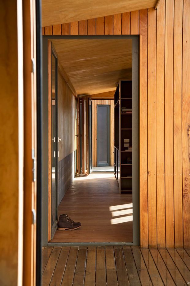 A warm, narrow timber-lined passage viewed through an open doorway. Vertical timber wall cladding continues across the ceiling and floor, with dark-framed glazed openings along the left side, built-in dark shelving on the right, and a doorway at the far end. Strong sunlight creates rectangular patches across the timber floor, while a pair of boots rests near the entrance.