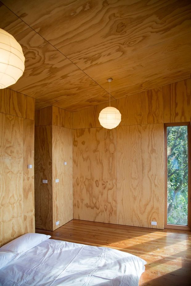 A warm, compact bedroom clad almost entirely in exposed plywood, including the walls and ceiling. A low bed with white bedding occupies the foreground, while spherical pendant lights, timber flooring, simple electrical outlets, and a tall timber-framed window create a minimal, cabin-like interior with strong natural light.