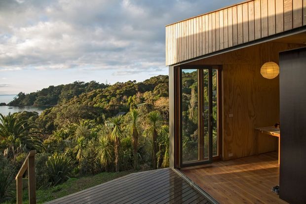 A modern timber-clad dwelling viewed from a covered deck, with an open glazed corner revealing a warm timber-lined interior and a compact kitchen area. The home overlooks dense native vegetation and a sheltered coastal or estuarine landscape. The building has a clean box-like form, vertical timber cladding, dark-framed glazing, and a low-pitched or flat roof edge.