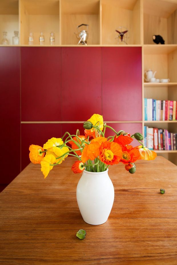 A warm interior vignette featuring a timber dining table with a white ceramic vase of bright orange and yellow flowers. Behind it is a full-height built-in timber shelving unit with open display compartments, books, glassware and decorative objects, contrasted by deep red cabinet fronts.