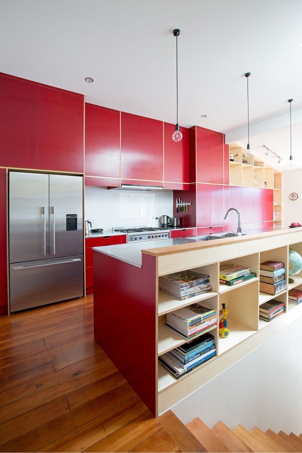 A bright contemporary kitchen viewed from a stairway, featuring vivid red full-height cabinetry with slim brass-toned trims, stainless-steel appliances, a white benchtop, timber flooring, and a large light-wood island with open shelving filled with books and objects. Pendant lights hang from a white ceiling, and timber stairs descend in the foreground.