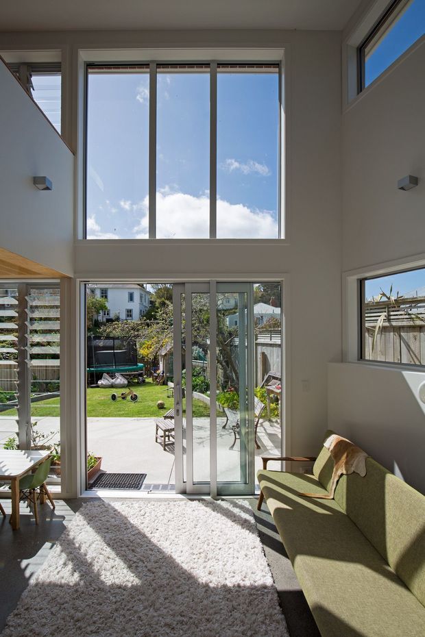 Bright contemporary living room with a double-height glazed wall, tall sliding glass doors, clerestory windows, and a view to a landscaped backyard. The room includes a green upholstered sofa, pale shag rug, timber dining furniture, and abundant natural light. A louvered window screen and exterior garden are visible beyond the glazing.