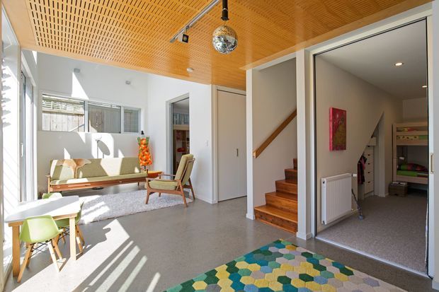 Bright, open-plan mid-century-inspired living area with large windows, a warm perforated timber ceiling, polished grey concrete flooring, and a colorful geometric rug. A compact lounge with vintage-style seating occupies the sunlit space, while an open doorway reveals a carpeted adjacent room and a timber staircase. Built-in storage and artwork add functional and decorative detail.