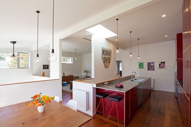 Open-plan kitchen and dining area with a large island featuring dark red cabinetry and a dark benchtop, timber flooring, white walls and ceilings, pendant lighting, skylight, and adjacent living space. A timber dining table occupies the foreground, with artwork and framed prints on the walls.