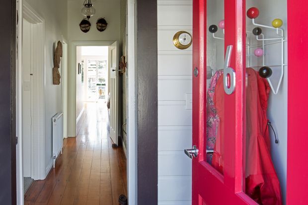 A narrow residential entryway and hallway viewed from the open front door. The vivid red glazed door dominates the right foreground, with a wall-mounted coat rack and hanging coats visible behind it. The hallway has warm timber floorboards, pale painted walls, white interior doors, a radiator, decorative wall objects, and daylight at the far end.