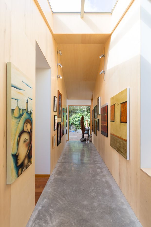 A long, narrow, gallery-like interior hallway with pale timber wall and ceiling panelling, a polished grey concrete floor, and a large skylight bringing in abundant natural light. Numerous framed artworks line both walls, with a glazed opening at the far end leading toward an outdoor area. Track spotlights provide additional illumination.