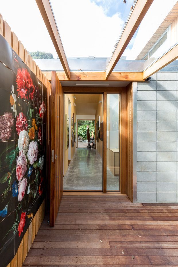 A bright, narrow courtyard-like entry passage with a timber deck, exposed timber beams, and a glazed overhead roof opening. The view looks through a timber-framed glass doorway into a long interior corridor. A large colourful floral mural lines the left wall, while the right side features a grey concrete block wall and upper glazing. The scene combines warm timber, natural light, and industrial masonry.