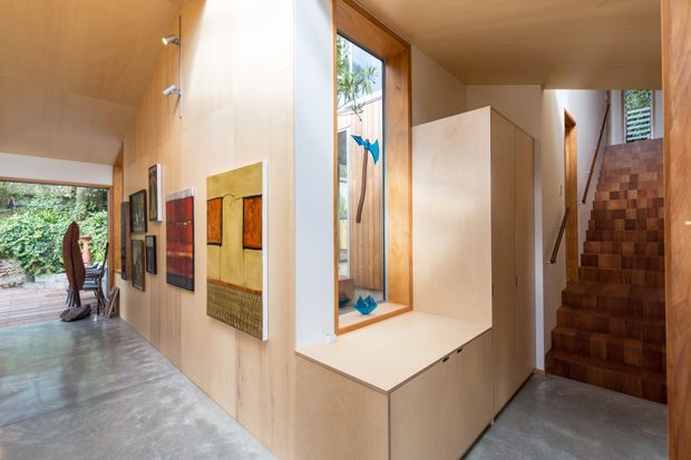 Warm contemporary interior corridor with a polished concrete floor, extensive light timber wall and ceiling lining, built-in timber cabinetry beneath a large internal window, and a timber stairway leading upward. Artwork and a desk area are visible along the open hallway, with greenery seen through glazing at the rear.