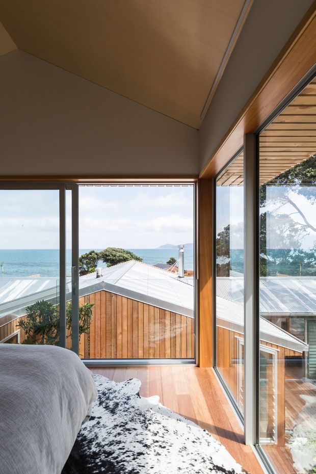 A bright bedroom corner with a bed in the foreground, warm timber flooring and extensive floor-to-ceiling glazing. The windows overlook a coastal setting with ocean views and neighbouring pitched metal roofs. Timber-framed glazing, exposed timber ceiling lining, and a sheltered timber-clad exterior are visible, creating a warm, contemporary coastal atmosphere.
