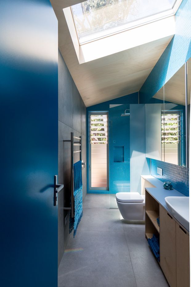 A bright, narrow contemporary bathroom viewed through a partially open deep-blue door. The room features vivid turquoise-blue mosaic wall tiles, a pale timber-lined sloping ceiling with a large skylight, grey concrete-look floor tiles, a wall-hung toilet, frameless glass shower enclosure, and a timber vanity with a white basin. Mirrored cabinetry or glazing reflects the vanity and louvred window, while natural light creates an airy, high-contrast atmosphere.