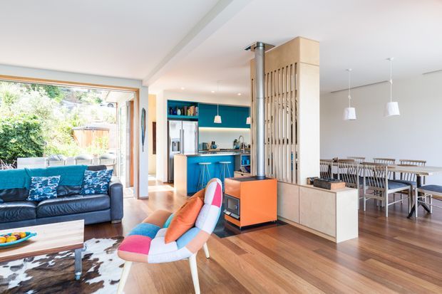 Open-plan residential interior showing a living room, kitchen and dining area. The space features warm timber flooring, white walls and ceiling, a bright turquoise kitchen with stainless-steel appliances, built-in timber cabinetry, a vertical timber screen dividing the zones, and colourful mid-century-inspired furniture. Large glazed doors connect the living area to greenery outside, creating a bright and casual family space.