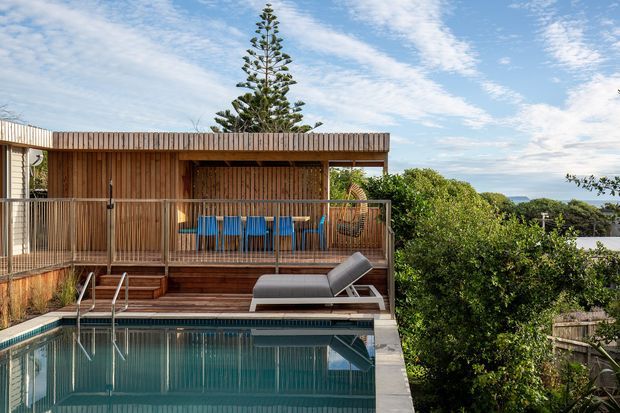 A modern timber-clad outdoor entertaining area beside a rectangular swimming pool. The single-storey structure has a flat roof, vertical timber screening and cladding, a covered dining deck with blue bar stools, and a timber balustrade. A poolside lounge chair and landscaped suburban surroundings are visible, with glimpses of a distant blue horizon.