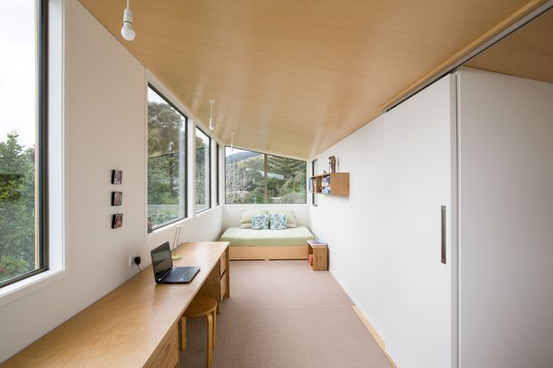 A bright, narrow modern interior combining a home office and compact lounge area. A long built-in timber desk runs along the window wall with a laptop and seating, while a pale upholstered daybed or sofa sits at the far end beneath large windows. White built-in storage occupies the right wall, and the ceiling is clad in warm timber. The glazing looks onto a leafy, mountainous landscape.