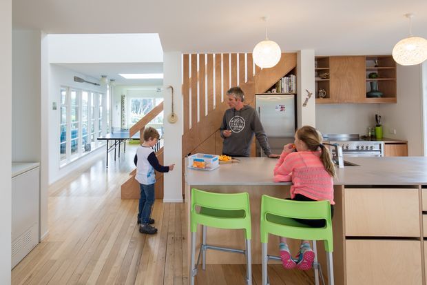 Bright open-plan kitchen and family living area with a large central island, pale timber flooring, light cabinetry, integrated appliances, and green children's chairs. A timber slat screen and stairway separate the kitchen from a light-filled hallway and adjoining family space. Two round pendant lights hang above the island.