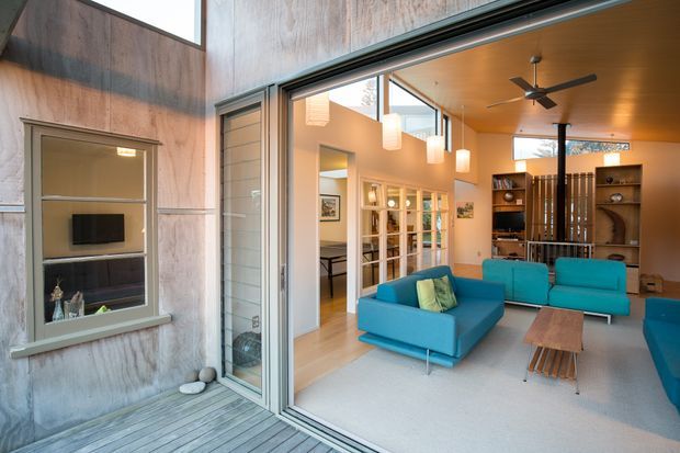 A contemporary open-plan living room viewed from a timber deck through large sliding glass doors. The room features vivid turquoise sofas, a pale area rug, a low timber coffee table, a warm timber ceiling, recessed and pendant lighting, a ceiling fan, built-in shelving, and a glazed internal partition. Textured concrete or plaster walls and extensive glazing create a strong indoor-outdoor connection.