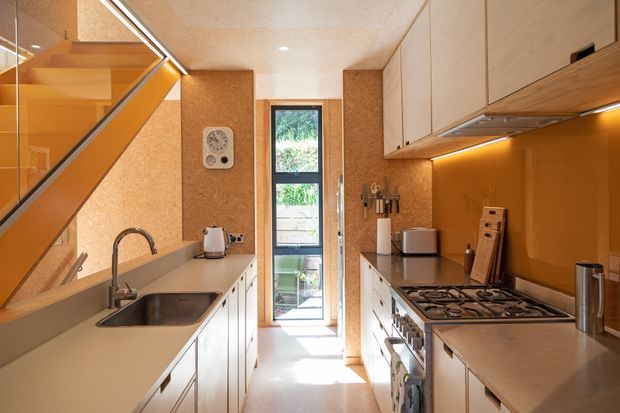 A narrow, warm-toned galley kitchen viewed toward a glazed rear door. Pale fixed cabinetry and light benchtops line both sides, with a stainless-steel sink and gooseneck tap on the left and a freestanding gas cooktop and oven on the right. An amber-toned splashback, cork-like wall panels, timber-framed glazing, and a compact stair with clear balustrade are visible. The kitchen is brightly daylit and efficiently arranged.