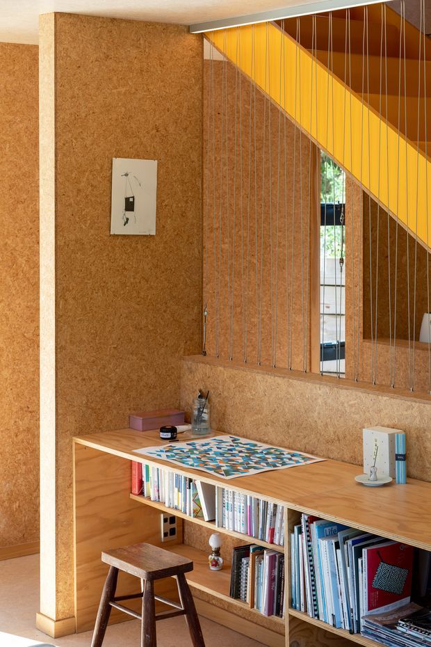 A warm, compact built-in study nook beneath a staircase. The space features extensive cork-textured wall and stair surfaces, open timber shelving filled with books, a timber desk, and a dark wooden stool. A striking yellow diagonal stair soffit and a screen of fine vertical metal rods separate the study from the adjoining area, with daylight visible beyond.