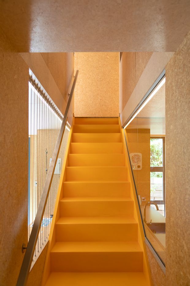 A narrow, vividly orange stairway rises through a tall interior void. The stairs and surrounding walls have a warm textured finish, with a slim metal handrail and vertical cable balustrade on the left. Full-height glazing and a large reflective glass panel on the right reveal glimpses of an adjacent interior and greenery outside. The composition is highly geometric, warm, and contemporary.