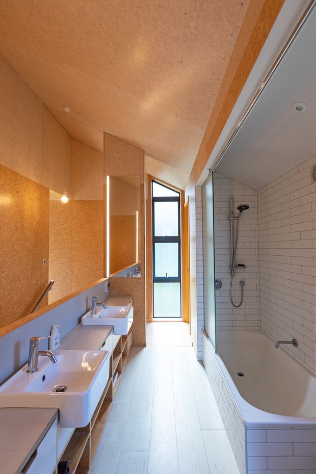 A narrow, elongated contemporary bathroom with two white vessel-style basins on a long vanity, open timber shelving, a large wall mirror, and warm cork-toned wall and ceiling panels. The opposite side contains a white tiled bathtub with overhead shower, handheld shower, and grab rail. A tall black-framed window at the end provides natural light, while pendant and recessed lighting illuminate the space.