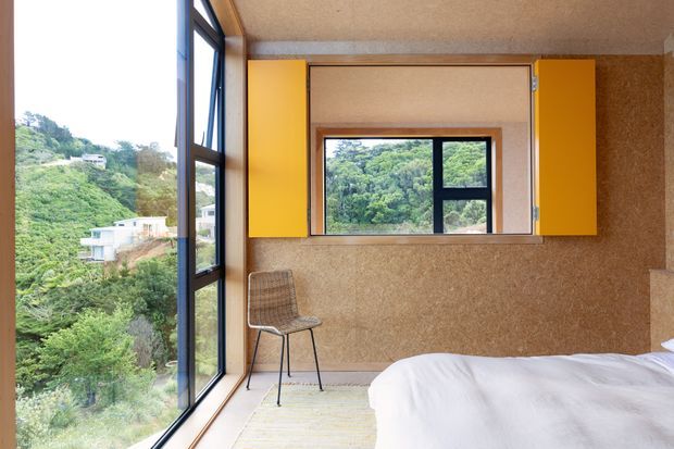 Compact bedroom with a partial view of a bed, a woven accent chair, full-height corner glazing, and a black-framed interior window. Warm cork-textured wall surfaces are paired with bright mustard-yellow built-in panels around the window. The room overlooks a lush, steeply vegetated hillside with scattered homes.