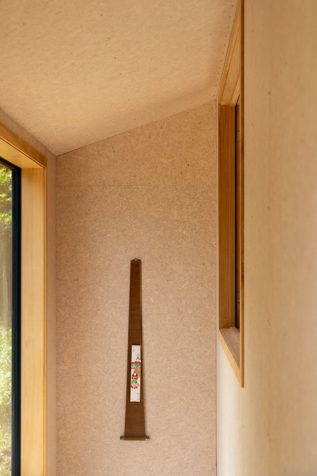 A narrow, warm-toned interior passage or entry area with plywood-lined walls and ceiling, timber-framed glazing on the left, and a deep timber-framed recessed opening on the right. A slim decorative wooden object is mounted vertically on the textured wall. The space has simple built-in detailing and natural timber finishes.