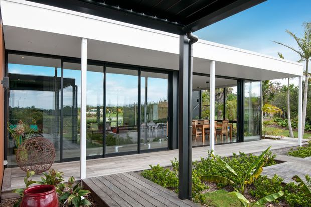 Contemporary single-storey home viewed from a covered timber deck and landscaped courtyard. The white flat-roofed building has extensive black-framed floor-to-ceiling glazing and sliding doors opening onto outdoor dining and living areas. Tropical planting surrounds the deck, while a dark metal pergola and downpipe frame the foreground.