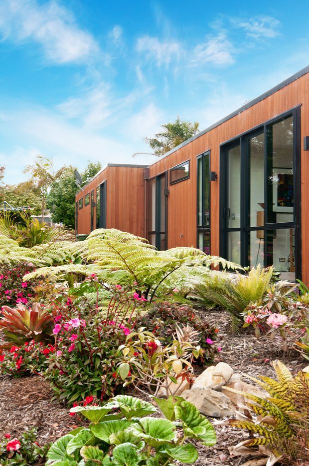 Exterior view of a low-profile contemporary building set within a lush landscaped garden. The facade features warm vertical timber cladding, dark metal window and door frames, extensive glazing, and a shallow sloping roofline. Ferns, flowering shrubs, tropical plants, mulch, and stone edging dominate the foreground, creating a vibrant garden setting.