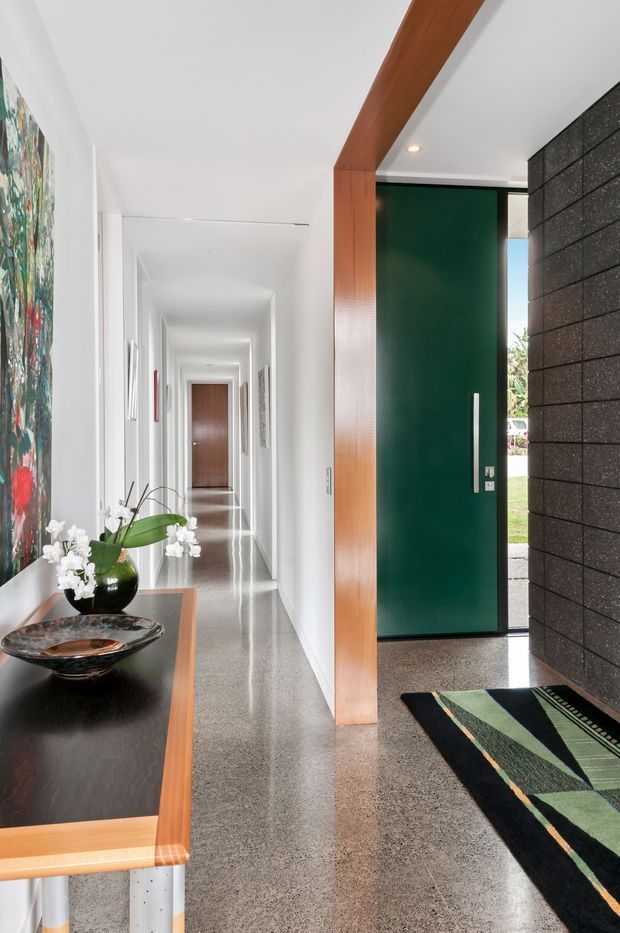 A contemporary residential entryway looking down a long, bright hallway. The space features polished grey terrazzo flooring, white walls, a bold dark green pivot front door, a charcoal textured masonry feature wall, and a warm timber structural beam and door surround. A slim console with a black surface, decorative bowl, orchid, and artwork adds colour and interest, while a patterned runner defines the entrance.