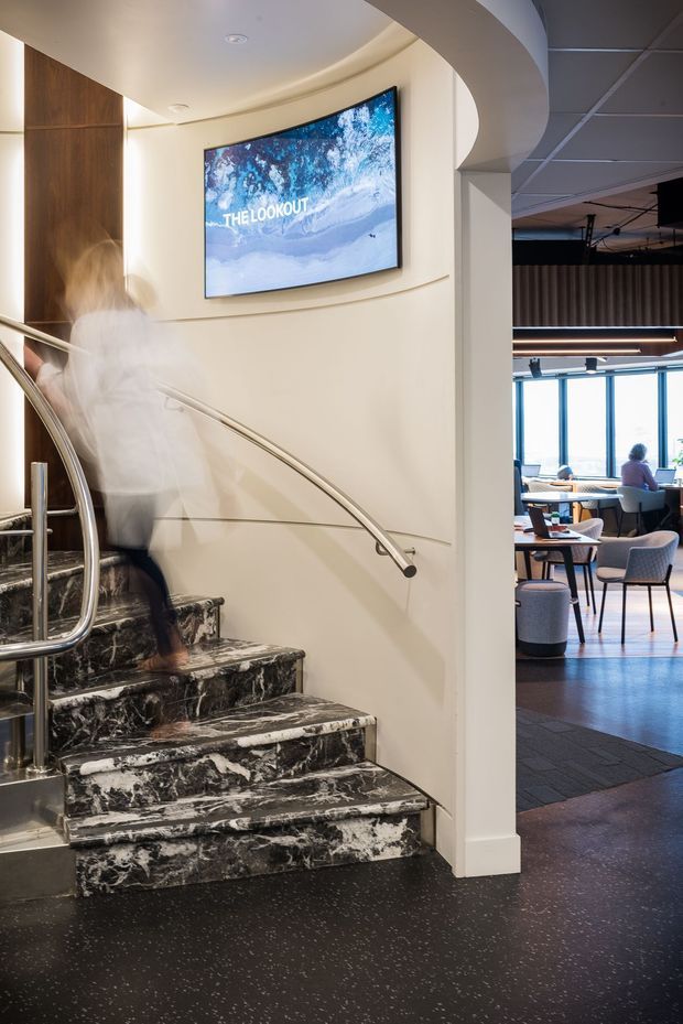 Interior view of a contemporary commercial space featuring a curved staircase with dramatic black-and-white marble steps, stainless steel handrails, warm cream walls, timber wall panelling, and an integrated wall-mounted digital display. An adjacent open seating and dining area is visible beyond the curved opening, with large windows and modern furniture.