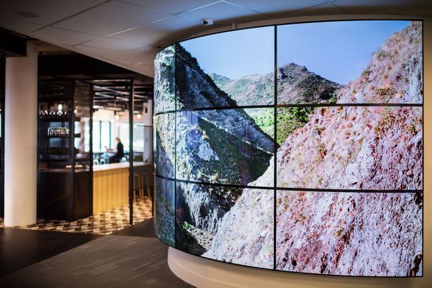 A contemporary commercial interior featuring a large curved video wall displaying a close-up mountain landscape. The surrounding space includes a bar or reception counter, dark shelving, patterned tiled flooring, carpeted circulation space, and a suspended acoustic tile ceiling. The dramatic digital installation is the dominant feature.