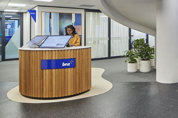 A contemporary commercial reception area featuring a curved, vertically ribbed timber reception desk with a blue BNZ sign, integrated display screens, grey carpet and speckled dark flooring. The space has large glazed partitions, a curved white architectural soffit and column, indoor plants, and abstract wall artwork.