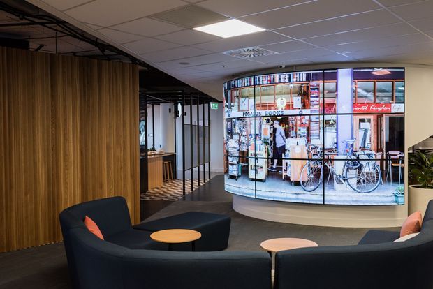 Contemporary commercial lounge or reception area with curved dark upholstered seating, small round tables, a tall timber slat feature wall, exposed dark ceiling services, and a large curved digital display showing a streetscape. Glazed partitions and a patterned carpeted floor add visual interest.