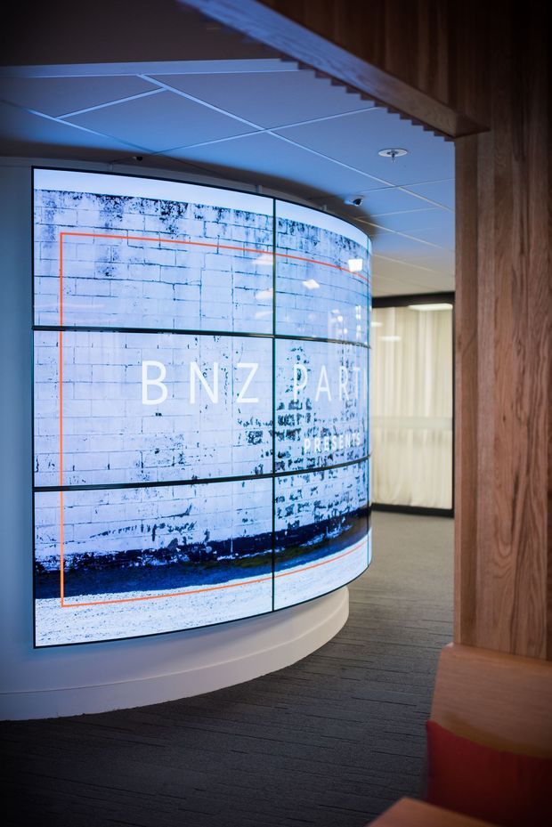 A contemporary commercial interior featuring a large curved digital video wall displaying BNZ branding and imagery. The space has a dark carpeted floor, recessed grid ceiling, warm vertical timber wall cladding, built-in timber seating with red upholstery, and a softly illuminated glazed or curtained area beyond.
