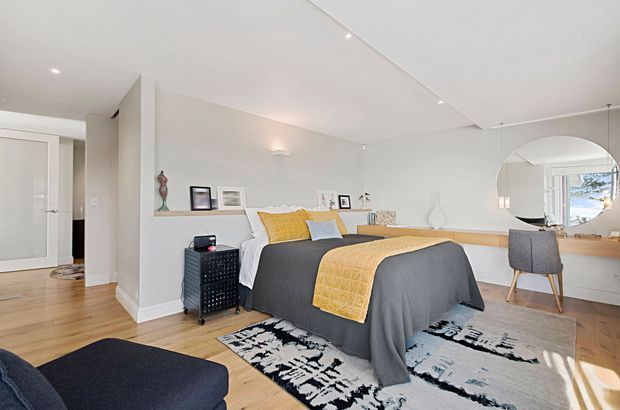 Bright, contemporary bedroom with a grey upholstered bed dressed in mustard-yellow and white bedding, set against pale walls and light timber flooring. A long built-in timber desk or shelf runs along the wall beneath a large round mirror, with a grey chair and minimal decorative objects. The room opens to a hallway through a frosted-glass door and includes a monochrome patterned rug and recessed ceiling lighting.