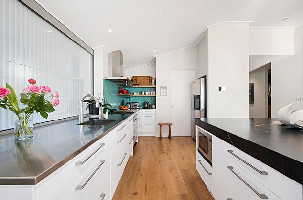 Bright contemporary kitchen with white cabinetry, long dark stone benchtops, timber flooring, a turquoise tiled splashback, open shelving, integrated appliances and a large window with vertical blinds. The space includes a central island and a peninsula-style work area, with white walls and ceiling creating an airy feel.