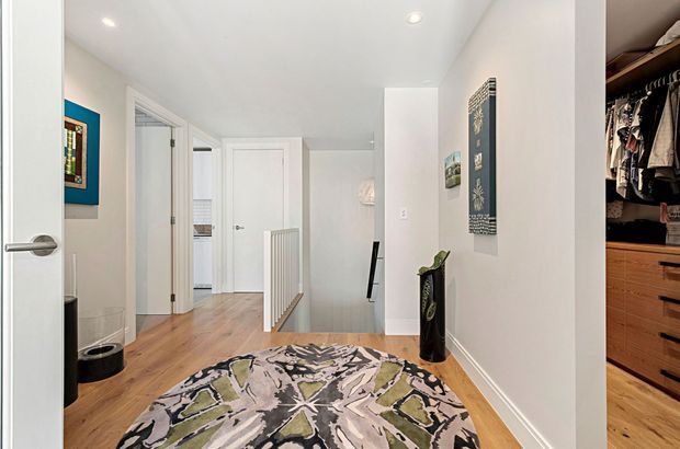 Bright contemporary hallway or landing with white walls, recessed ceiling lights, warm timber flooring, and a patterned area rug. Multiple white doors open off the hall, with a stair railing visible ahead and a walk-in wardrobe or storage area visible at right.