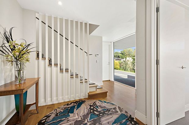 Bright contemporary entry foyer with a timber staircase screened by vertical white slats, warm timber flooring, a patterned area rug, and a small console table with floral arrangement. An open front door provides a view toward the street and landscaped exterior.